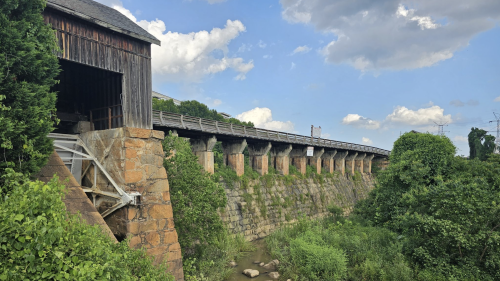 A stone bridge with a wooden structure above, surrounded by lush greenery and a small stream below.