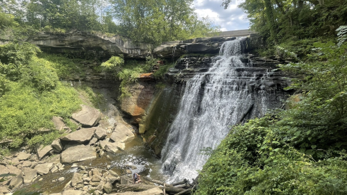 A scenic waterfall cascades over rocky cliffs, surrounded by lush greenery and a bright blue sky.