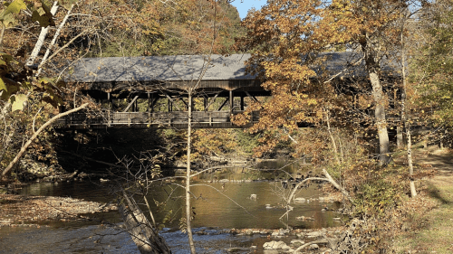 A covered bridge spans a serene river, surrounded by autumn foliage and trees reflecting in the water.