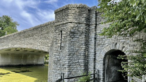 A stone bridge with an archway, surrounded by greenery and reflecting water, under a blue sky with clouds.