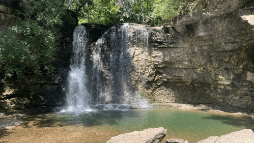 A serene waterfall cascading into a clear pool, surrounded by lush greenery and rocky cliffs.