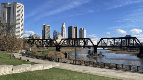 A view of a city skyline with a bridge over a river, surrounded by green grass and blue skies.