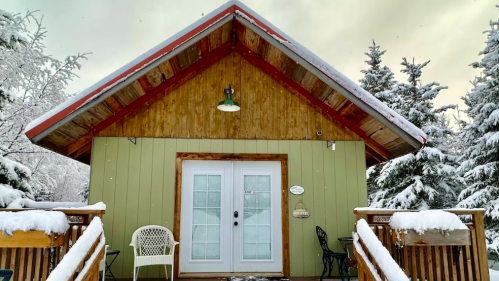 A cozy cabin with a wooden deck, surrounded by snow-covered trees under a cloudy sky.