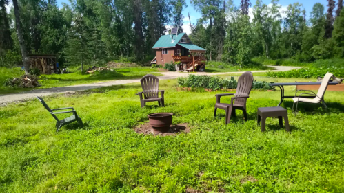A grassy area with several chairs arranged around a fire pit, surrounded by trees and a house in the background.
