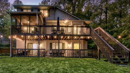 A two-story house with a deck, surrounded by trees, illuminated by string lights at dusk. Green lawn in the foreground.