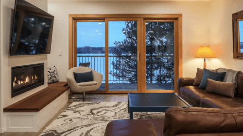 Cozy living room with a fireplace, large windows overlooking a lake, and modern furniture in warm tones.