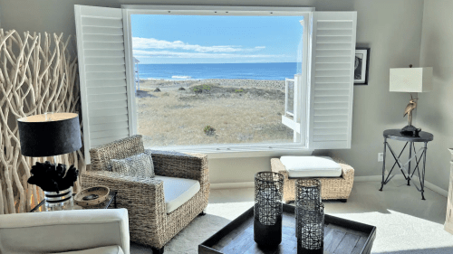 Cozy living room with a view of the ocean through large windows, featuring wicker furniture and a stylish lamp.