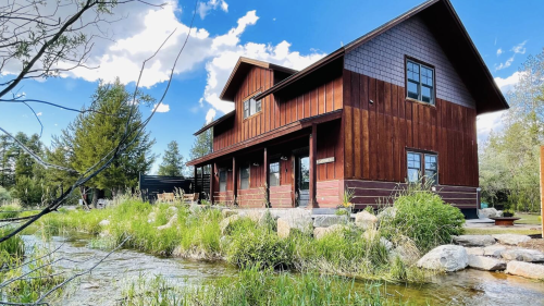 A rustic wooden house beside a stream, surrounded by greenery and under a blue sky with scattered clouds.