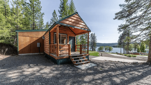 A cozy wooden cabin with a porch, surrounded by trees and overlooking a lake in the background.
