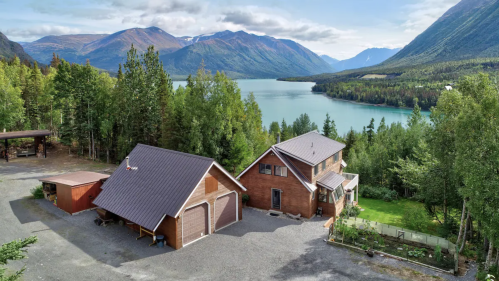 Aerial view of a wooden house and garage by a lake, surrounded by trees and mountains under a blue sky.