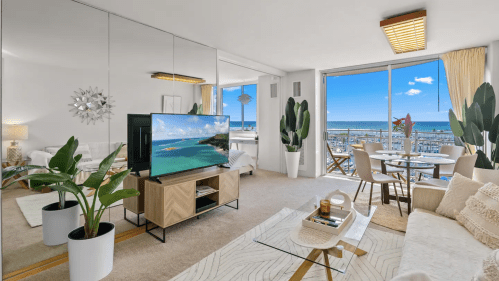 Modern living room with a large TV, plants, and a view of the ocean through a glass door. Bright and airy atmosphere.