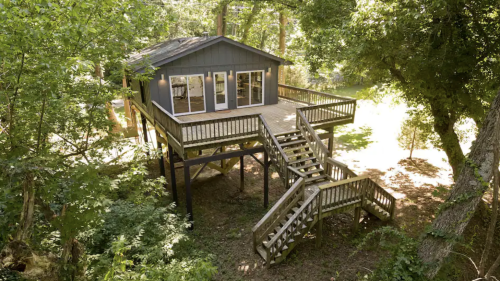 A modern cabin on stilts surrounded by trees, featuring a large deck and staircase leading to the ground.
