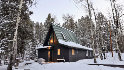 A cozy A-frame cabin surrounded by snow-covered trees, with warm light glowing from the windows at dusk.