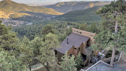 A modern house nestled among trees, overlooking a mountainous landscape with rolling hills and a clear sky.