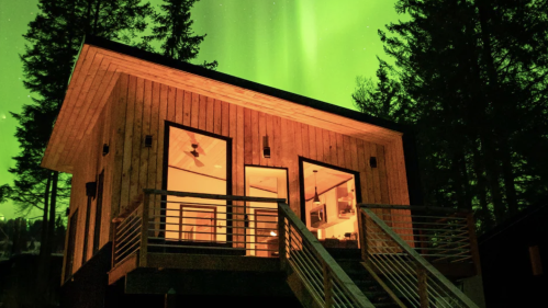A cozy wooden cabin illuminated by the northern lights, surrounded by tall trees at night.