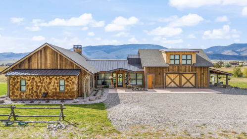 A spacious wooden house with a modern design, surrounded by grass and mountains under a blue sky.