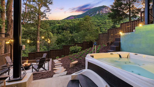 A scenic view of a hot tub overlooking a lush mountain landscape at sunset, with outdoor seating and string lights.
