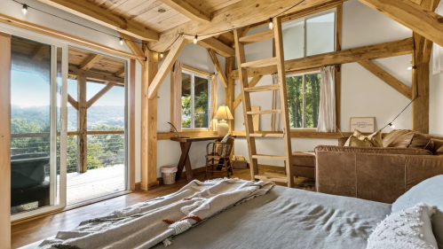 Cozy wooden cabin interior with a bed, desk, and large windows showcasing a scenic view of nature.
