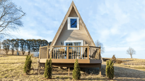 A cozy A-frame house with a wooden deck, surrounded by grass and small trees under a clear blue sky.