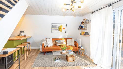 Bright living room with an orange sofa, wooden bar stools, and a modern light fixture, featuring a cozy, inviting atmosphere.