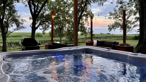 A hot tub on a patio surrounded by trees, with string lights and a view of a sunset over a vineyard.