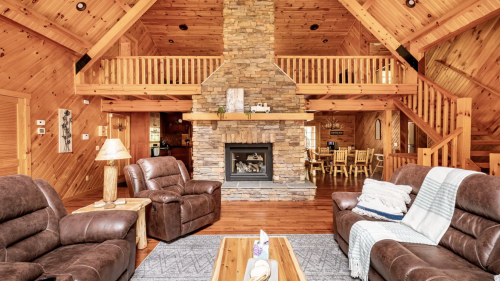 Cozy wooden cabin interior with a stone fireplace, leather sofas, and a dining area in a warm, inviting atmosphere.