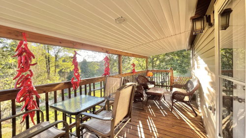 A cozy porch with a table and chairs, decorated with red chili peppers, surrounded by greenery and sunlight.