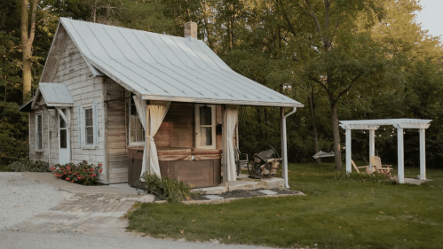A quaint wooden cottage with a metal roof, surrounded by greenery and a small patio area with chairs.