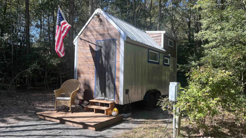 A tiny house on wheels with a wooden deck, an American flag, and a chair, surrounded by trees in a wooded area.