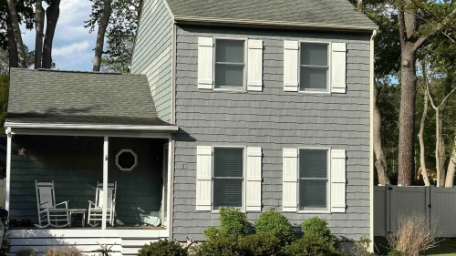 A two-story gray house with white shutters and a porch, surrounded by greenery and trees.