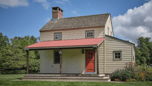 A charming two-story house with a red door and roof, surrounded by green grass and trees under a blue sky.