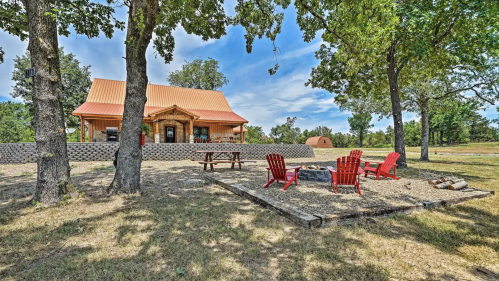 A cozy cabin with a red roof surrounded by trees, featuring a fire pit and picnic area with red chairs.