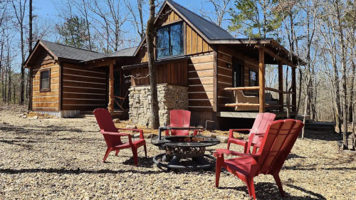 A cozy wooden cabin surrounded by trees, featuring a fire pit and red chairs in a gravel area.