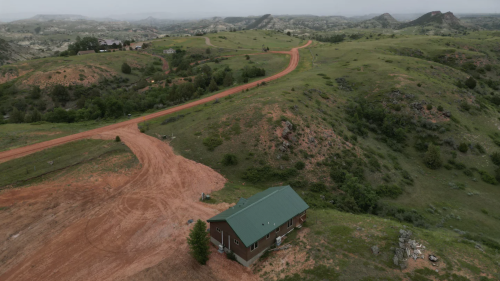 Aerial view of a green-roofed house on a red dirt road, surrounded by rolling hills and overcast skies.