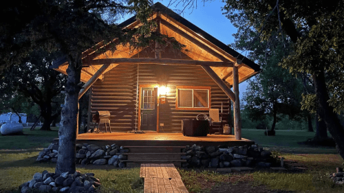 A cozy log cabin illuminated at dusk, surrounded by trees and a wooden walkway leading to the entrance.