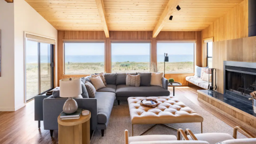 Modern living room with wooden beams, large windows, and a cozy seating area overlooking a grassy landscape.