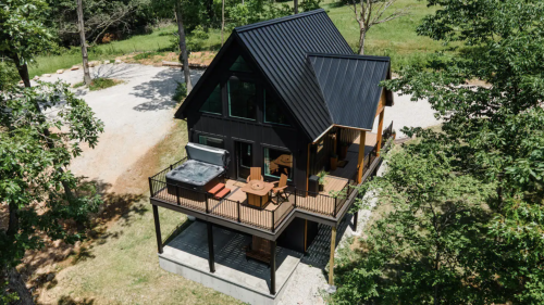 A modern black house with a metal roof, surrounded by trees and a grassy area, featuring a deck and outdoor seating.