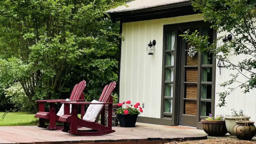 A cozy house with a wooden deck, two red chairs, and lush greenery surrounding it.