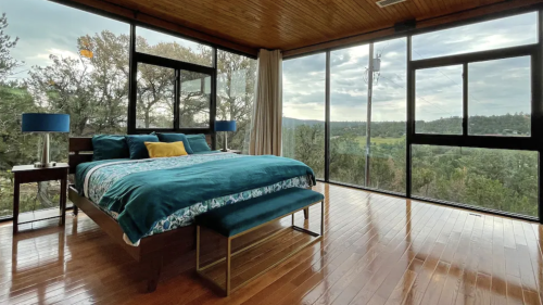Bright bedroom with large windows, wooden ceiling, and a bed dressed in teal linens, overlooking a scenic landscape.