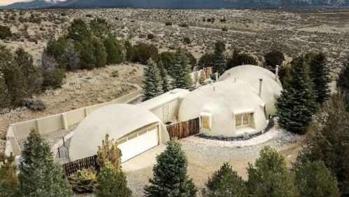 A unique dome-shaped house surrounded by trees and mountains, set in a vast, open landscape at sunset.
