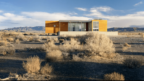 Modern wooden house in a barren landscape with sparse vegetation and mountains in the background under a clear sky.