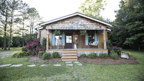 A charming wooden cottage with a shingled roof, surrounded by colorful flowers and greenery, on a sunny day.