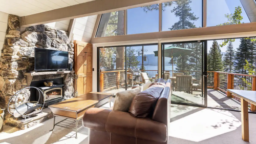 Cozy living room with stone fireplace, large windows, and a view of trees and a lake outside.