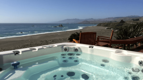 A hot tub overlooking a beach and ocean, with a clear blue sky and distant hills in the background.