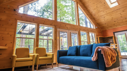 Cozy interior of a cabin with large windows, a blue sofa, and yellow chairs, surrounded by trees.
