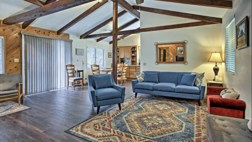 Cozy living room with wooden beams, blue sofa, armchair, and a patterned rug, featuring a dining area in the background.