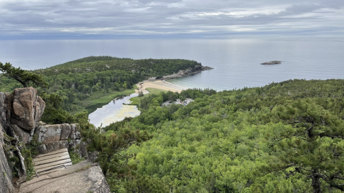 A scenic view of a coastal landscape with lush greenery, a sandy beach, and calm waters under a cloudy sky.