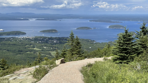 A scenic view from a mountain trail overlooking a blue bay with islands and lush greenery under a partly cloudy sky.