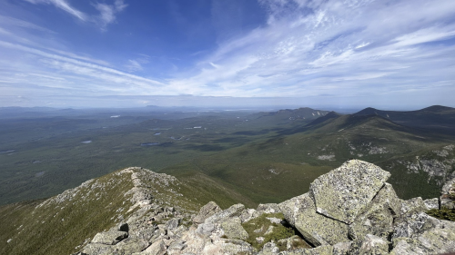 A panoramic view from a mountain peak, showcasing lush green valleys and distant mountains under a blue sky.