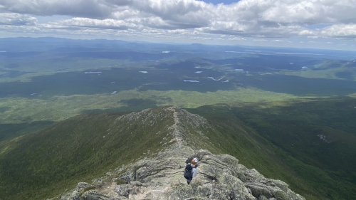 A person climbs rocky terrain at the summit of a mountain, overlooking a vast green landscape and cloudy sky.